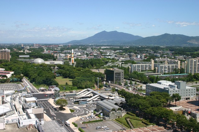 手前にはバスターミナルやたくさんのビルがあり、奥には山並みが見える様子を高い場所から撮った写真