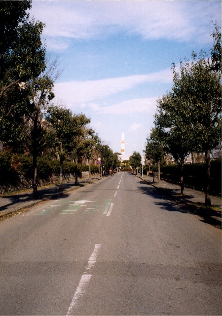 街路樹が立ち並ぶ直線道路の先に白と赤のボーダー色のロケットが立っている写真