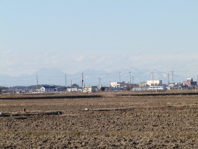 作物が収穫された後の畑が広がり、民家越しに見える日光連山の写真