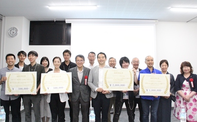 採択決定書のボードを持っている4団体の代表のメンバーが笑顔で2列に並んでいる記念写真