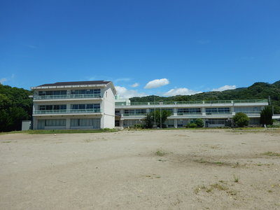 周囲を低い山に囲まれた2階建てと3階建ての旧小田小学校を運動場から撮影した写真