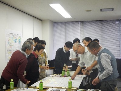 テーブルに置かれた資料を四方から囲んで眺めている関係者たちが立って協議している写真