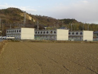奥の小高い山の手前に3棟建つ2階建ての小田第二団地を隣接する畑側から撮影した写真