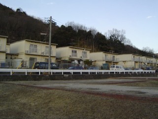 木が生い茂っている小高い山の麓に6棟建ち並んでいる2階建ての北条日向団地を道路を挟んだ反対側の敷地から撮影した写真