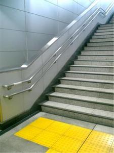 駅内にある階段の写真。壁には金属製の手すりが二つ取付られており、のぼりはじめの地面の部分には黄色い点字タイルが設置されている。