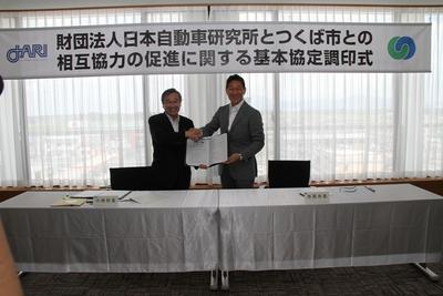 「一般財団法人日本自動車研究所とつくば市の相互協力の促進に関する基本協定について」と書かれた幕が張られ、その前で協定書を持ち握手をしているJARI所長と市原市長の写真