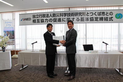 協定書を開いて一緒に持って立ち、握手をしている岡田義光理事長と市長の写真