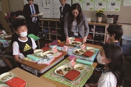 スーツ姿の女性と小学生たちが給食の乗った机をくっ付けて座る写真