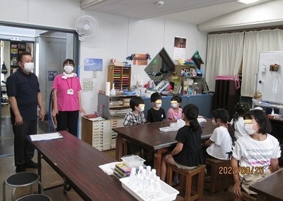 実験隊の様子で、テーブルの上にプラスチック製品や工作道具が置いてあり、こども達に説明をしている写真