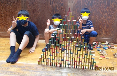 積み上げたドミノと一緒にピースをする3人の男の子の写真