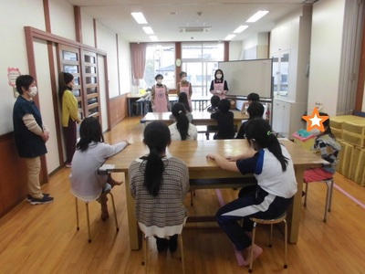 ピンク色のエプロンを着用した3名の女性スタッフの話を大きな2つのテーブルを囲んで座って聞いている子供たちの後ろ姿の写真