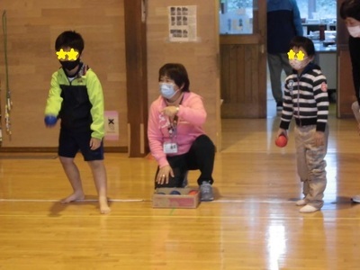 青いボールを持った子供と赤いボールを持った子供、ボールの入った箱の前に先生がいる様子の写真