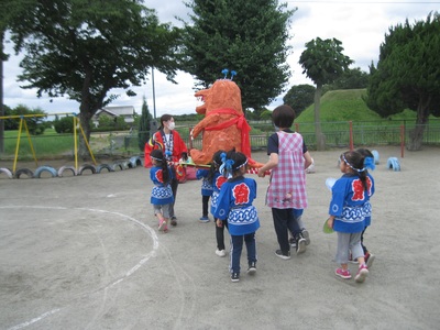 オレンジ色の体の怪獣の様な形をした神輿を先生と子供たちが担いでいる写真