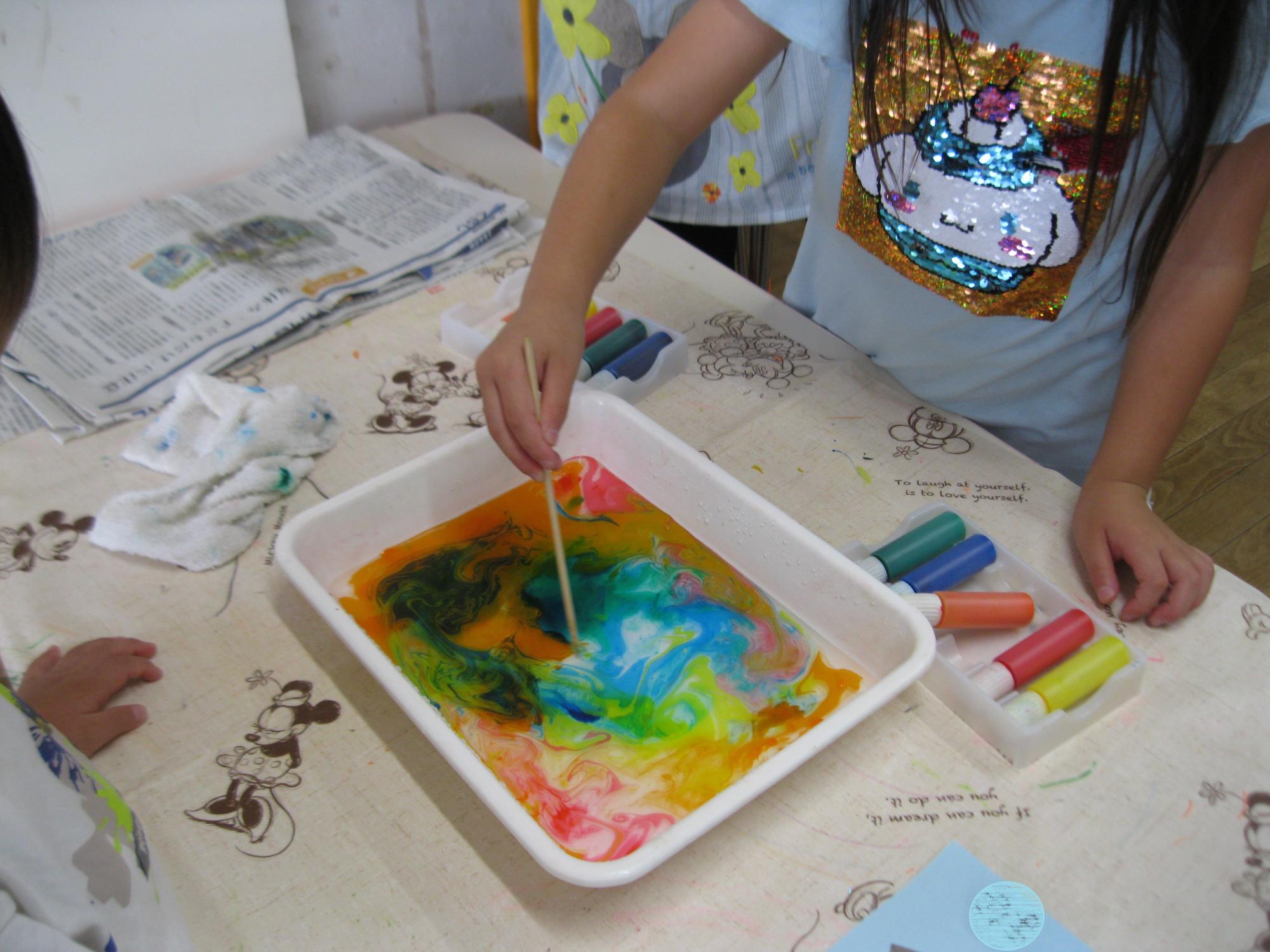 5歳児がマーブリング用の絵の具をカラフルに水の入ってるケースへ垂らし棒でやさしく混ぜています。