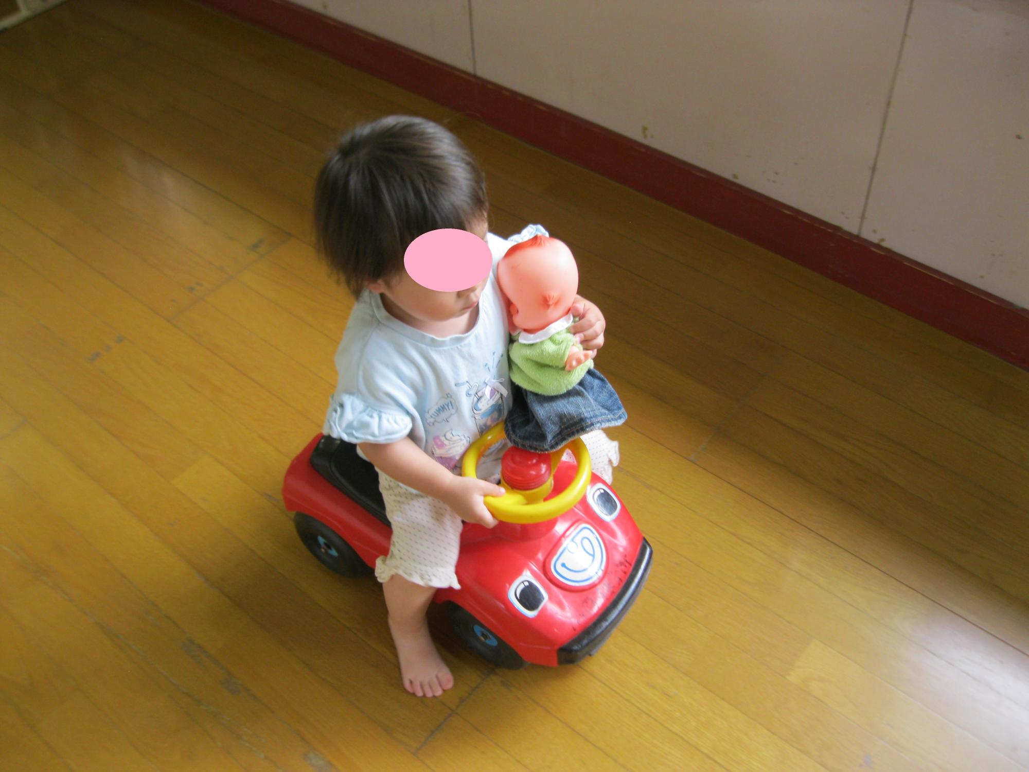1歳児の女の子がキューピーを片手にもって車ののりものに乗っています。