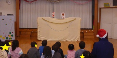大きな布が掛けられた上でサンタクロースとトナカイの紙人形劇が行われているのを床に座って熱心に見ている子供たちの写真