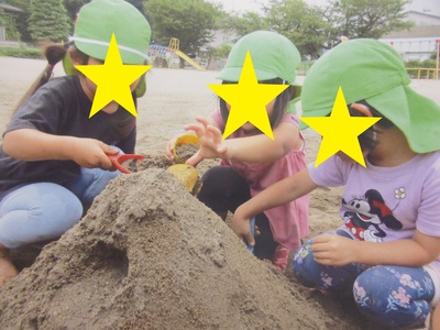 緑色の帽子を被った3名の女の子が砂場で山の形を作っている砂遊びの様子の写真