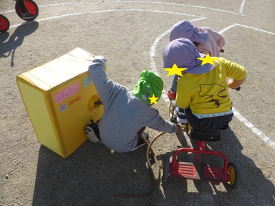 三輪車に乗った紫色の帽子を被った2名の園児と緑色の帽子を被った園児がガソリンスタンド遊びをしている様子の写真