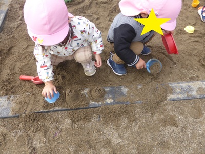 砂場で容器に入れた砂をひっくり返してカップケーキを作って遊んでいる2名の園児の写真