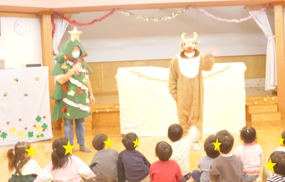 床に座っている子供たちの前でクリスマスツリーの着ぐるみを着た先生ががマイクを持ち、トナカイの着ぐるみを着た先生が子供たちに向かって手を振っている写真