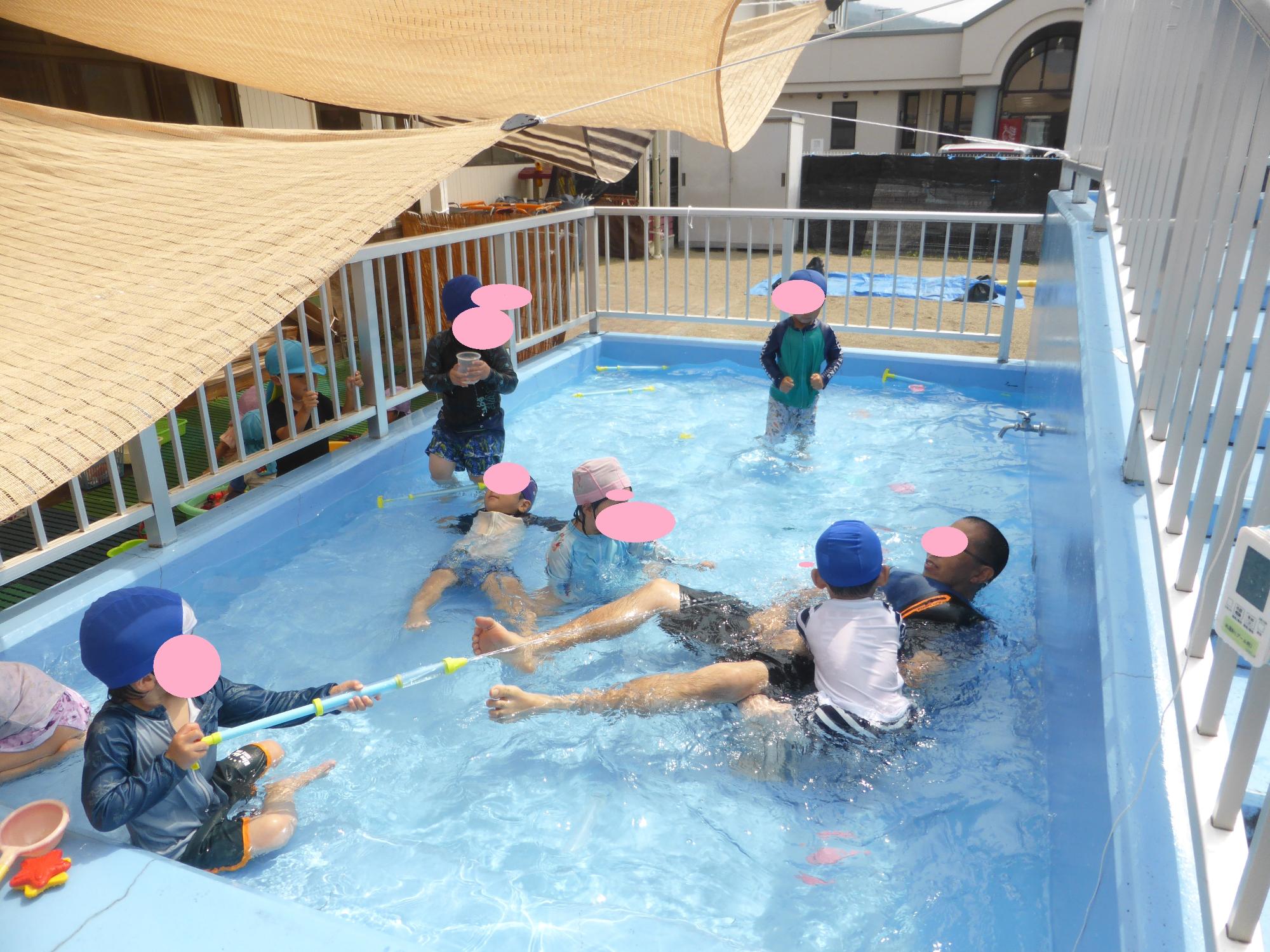 3歳児の子どもたちが、担任と一緒になってプールで遊んでいます。水鉄砲で担任を狙っています。