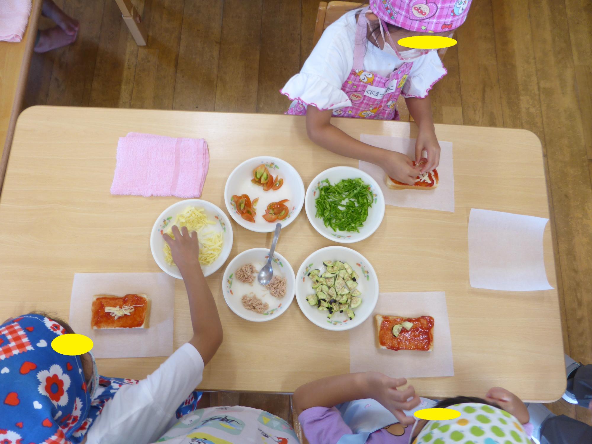 子どもたちが、ピーマン、トマト、ナス、ツナ、チーズをパンにのせて、ピザづくりをしています。