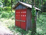 通路沿いに立つ1本の木の傍に設置された赤色の扉に白文字で消防団のポンプ格納庫と書かれている小さな小屋の写真