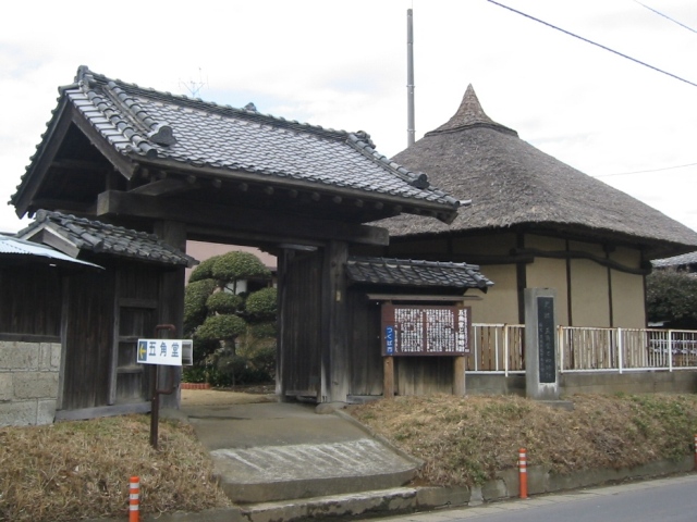 瓦屋根の門と茅葺き屋根の建物が並んでおり、門の前に「五角堂」という看板が立てられている写真