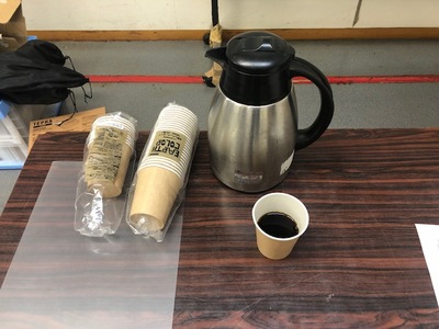 テーブル上に、袋に入った未使用の紙コップとポットが置かれており、一つだけ出されている紙コップにはコーヒーが入っている写真