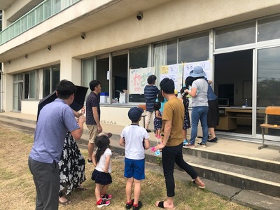 教室の外に張り出されている紙の前で、子どもを連れた地域の参加者たちが並んでいる報告会の様子の写真