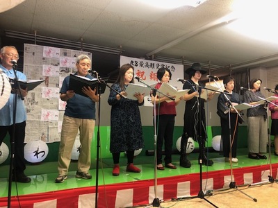小さなステージ設置されたマイクスタンドの前に立った8名の出演者が朗読劇を行っている様子の写真