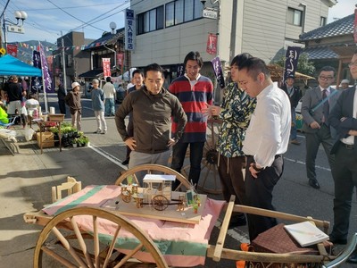 布が掛けられた大八車の上に小さな家具を乗せたミニチュアの大八車が展示されている前で見ている人達に男性が説明をしている写真