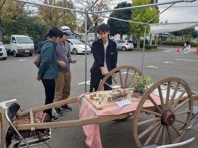 男女の見学者に大八車の上に乗せているミニチュアの展示物の説明を行っている男性の写真