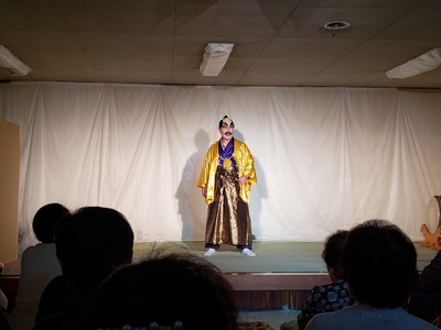 ちょんまげのカツラを被り黄色の着物を着た男性がステージに立っている演劇のワンシーンの写真