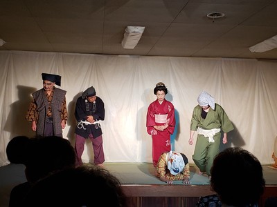 時代劇の装いをした5名の出演者がステージに立ち、床に両手をついて頭を下げている女性を他の出演者が見ている「あっぱれ伊賀七」公演のワンシーンの写真