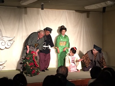 時代劇の装いをした5名の役者がステージに立ち、クリスマスツリーを持っている左側の男性を右側に座っている男性と女性が見ているシーンの写真