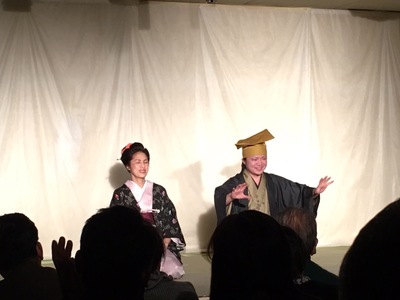 舞台の上に時代劇の装いをした女性と男性が正座し、右側の男性が両手を前に出し広げている演劇のワンシーンの写真