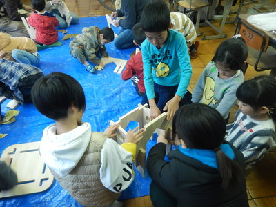 室内の床に敷かれたビニールシートの上で子供たちが力を合わせて木材を組み立てているものづくりワークショップの様子の写真