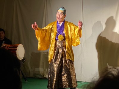 ちょんまげのカツラを被り黄色の着物を着た男性が両手を広げて話をしている演劇のワンシーンの写真