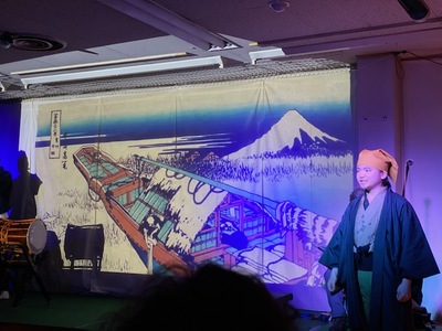 雪が積もった富士山の手前に猪牙船が描かれた絵がスクリーンに映し出された右側に着物を着た男性が立っている伊賀七BRAND DAYの様子の写真