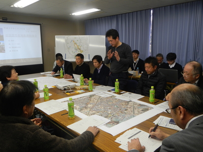 長机に様々な資料が置かれた部屋で、資料を見ながら話をしている一人の男性と、椅子に座って男性の話を聞いている関係者たちの写真