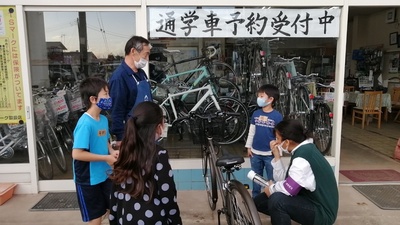 沢山の自転車が陳列されている「通学者予約受付中」と大文字で書かれた紙が貼られたガラス窓の店舗の前で1台の自転車を囲んで店主の男性と子ども達が話をしている職場体験の様子の写真
