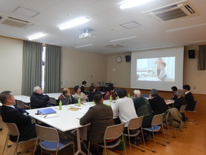 白いテーブルが並べられた室内で、プロジェクタースクリーンに映し出された映像を見ている参加者たちの写真