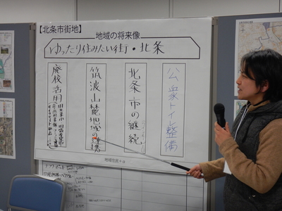 左手にマイク、右手に指示棒を持った男性が貼られた模造紙の右側に立ち発表を行っている様子の写真