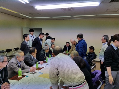 資料が置かれた机を囲んで座っている参加者が右奥に立っている男性の話を聞いている勉強会の様子の写真