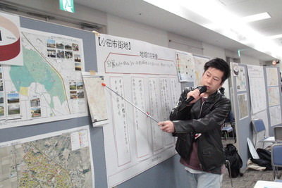 男性が右手でマイクを持ち、左手で指示棒を持って掲示板に貼られた地図を差している発表の様子の写真