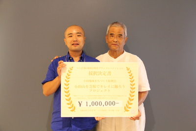 小田地域まちづくり振興会の2名の男性が一緒に採択決定書のボードを持っている写真