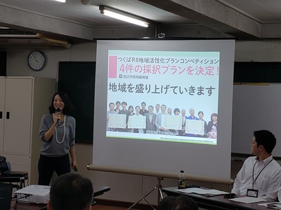 スクリーンに「地域を盛り上げていきます」の文言が映しだされており、スクリーン横で藤田教授がマイクを手に持って話をしている写真