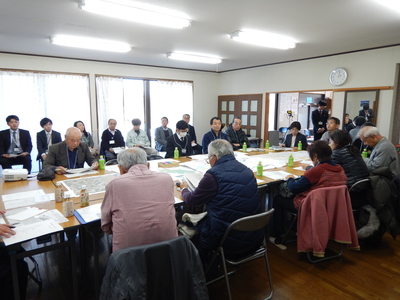 長机が並べられた部屋で、椅子に座り向かい合って資料を確認している参加者たちと、窓側に設置された椅子に座って参加者の様子を見ている関係者たちの写真