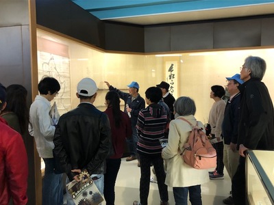 資料館で一つの展示物を説明している男性と、男性の話を聞いているツアー参加者たちの写真